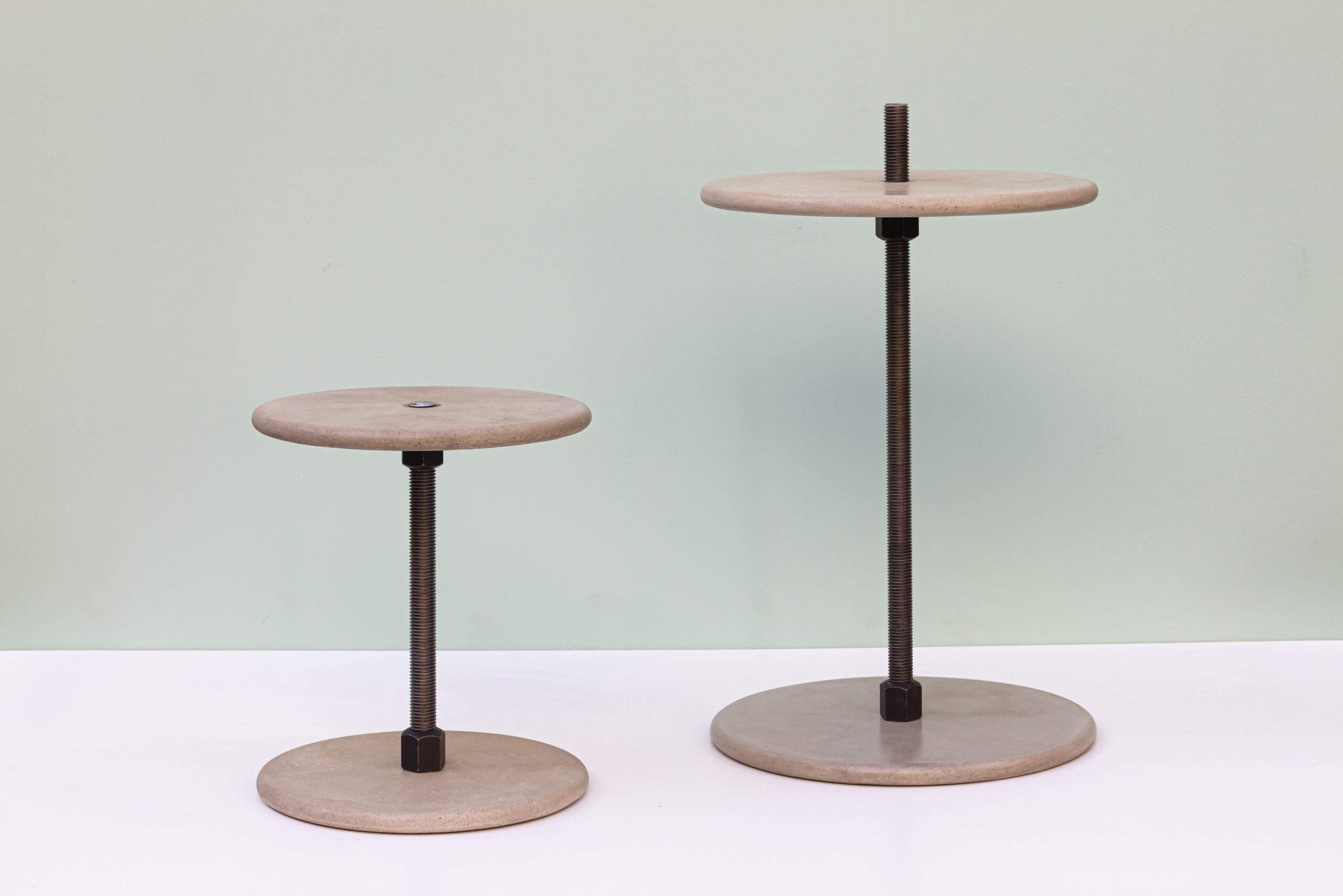 Ich sehe zwei minimalistische Betontische auf weißer Fläche vor einem hellgrünen Hintergrund. Beide haben runde Betonplatten oben und unten, verbunden durch eine zentrale, sichtbare Gewindestange aus dunklem Stahl; der rechte ist höher als der linke. Die Wirkung ist sehr ruhig und skulptural: klare Geometrie, rohe Materialien, viel Luft im Bild. Wenn du willst, kann ich dir daraus direkt einen kurzen Insta-Text machen, der genau zu diesem Foto passt.