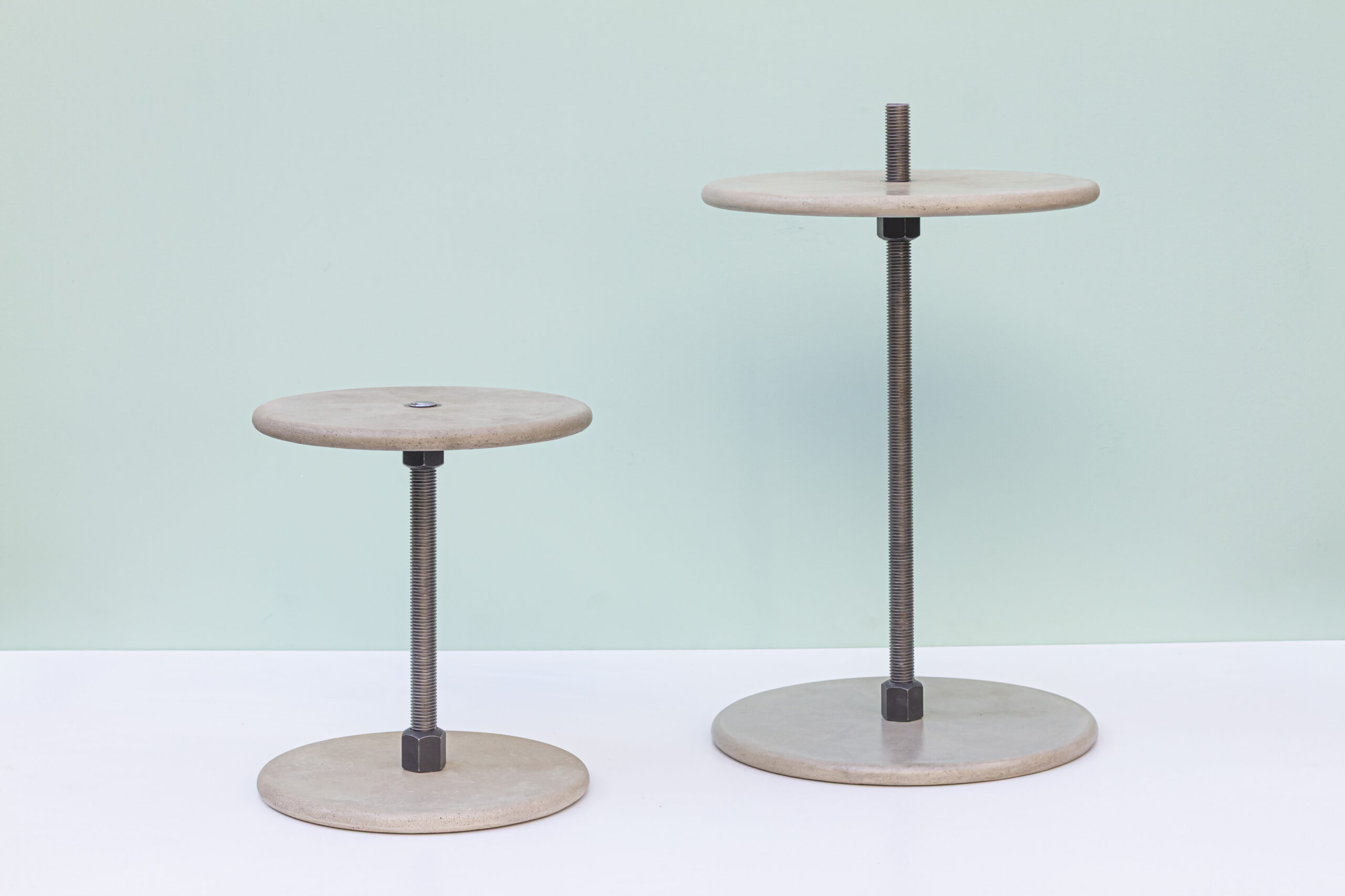zwei minimalistische Betontische auf weißer Fläche vor einem hellgrünen Hintergrund. Beide haben runde Betonplatten oben und unten, verbunden durch eine zentrale, sichtbare Gewindestange aus dunklem Stahl; der rechte ist höher als der linke. Die Wirkung ist sehr ruhig und skulptural: klare Geometrie, rohe Materialien, viel Luft im Bild.